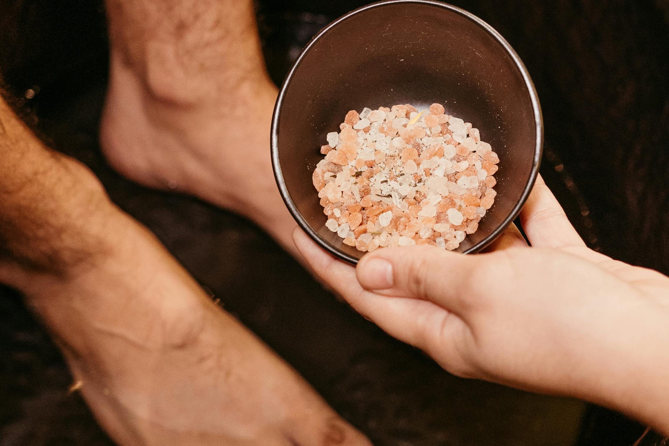 Une main tenant un bol contenant des cristaux de sel, tandis que des pieds nus sont visibles au fond.